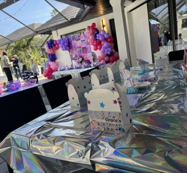 The outdoor birthday celebration is set with a charming movie-themed ambiance, featuring a table elegantly draped in a shiny silver foil cover reminiscent of a classic Movie Concession Stand Rental. The centerpiece is surrounded by vibrantly wrapped gift boxes and an array of cheerful, colorful balloons that add a festive and lively backdrop, perfect for any cinematic-inspired birthday bash.