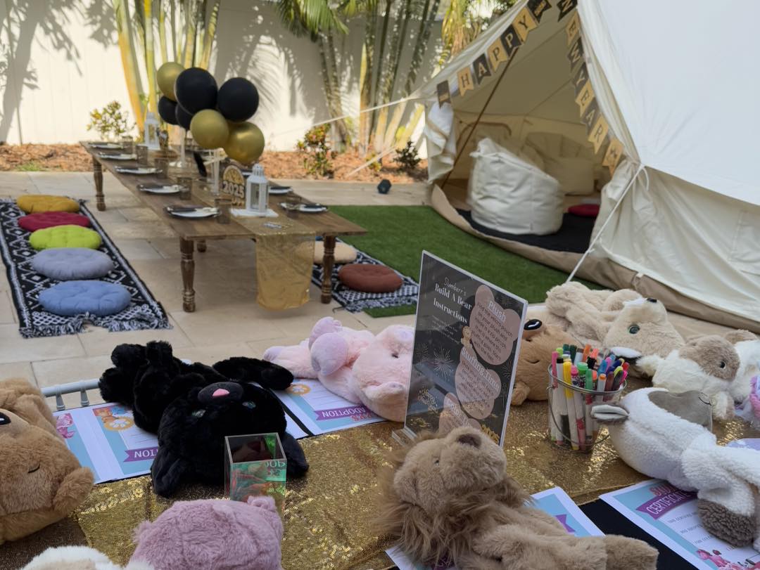 Celebrate in style with this enchanting outdoor birthday party setup that promises fun and creativity for kids. Set amidst lush greenery, a charming white tent provides a cozy haven filled with colorful cushions and a rustic low table adorned with an array of plush stuffed animals and vibrant art supplies. Adding to the festive ambiance, eye-catching black and gold balloons float gracefully above, exuding elegance and flair. The pièce de résistance is the Movie Concession Stand Rental, transforming the gathering into a Hollywood-themed extravaganza with its glamorous touch. Ideal for families looking to create magical memories, this delightful celebration is both imaginative and picture-perfect for creating lasting impressions.