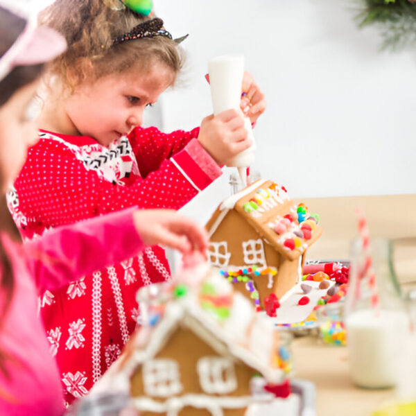 Two joyful children immerse themselves in the festive spirit as they craft gingerbread houses at a beautifully adorned holiday table. One child expertly handles a piping bag, adding intricate icing designs, while the other meticulously places colorful candy decorations. The scene captures warm holiday cheer and creativity, with gingerbread house decorating ideas and family bonding at its heart.