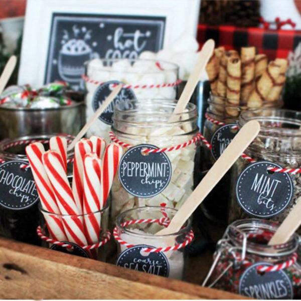 A delightful arrangement of mason jars on a rustic wooden table, each brimming with festive ingredients perfect for holiday indulgence: candy canes, rich chocolate chips, refreshing peppermint chips, vibrant mint bits, and colorful sprinkles. Each jar is accompanied by a charming wooden spoon and an elegant label that adds to the cozy aesthetic. In the background, a charming "Hot Cocoa" sign invites you to create your own warm winter beverage masterpiece. This enticing display not only evokes warmth and cheer but also optimizes your festive gathering with its ready-to-use cocoa toppings. Perfect for those seeking inspiration for holiday-themed DIY cocoa bars or creative gift ideas this season!