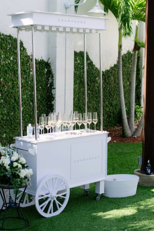 Elegant white champagne cart adorned with sparkling glasses and vibrant flowers, set amidst lush greenery on a sunlit grassy area beside a verdant wall. Ideal for weddings or outdoor celebrations, this picturesque setting combines nature and sophistication in perfect harmony.