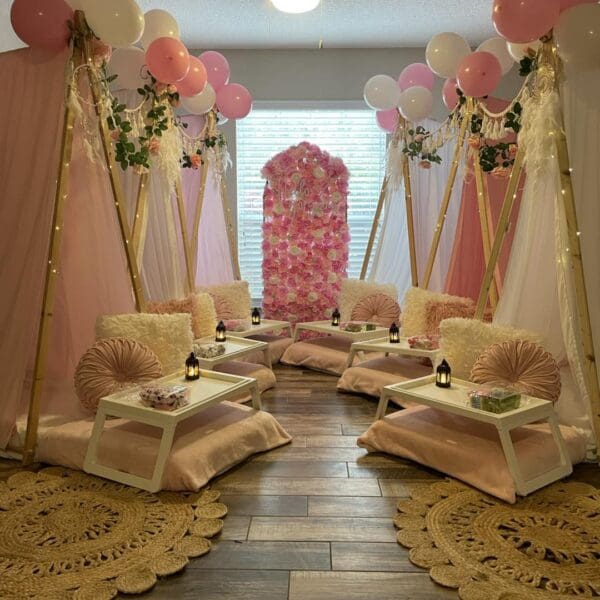 Immerse yourself in a whimsical indoor oasis with this cozy setup, perfect for a magical movie night experience. Delight in the enchanting pink and white teepee tents surrounded by plush cushions and elegant low tables, creating an intimate setting. The scene is adorned with vibrant balloons and lush floral decor, adding a splash of color and charm. An inflatable projector screen hints at a playful outdoor cinema vibe, while soft lighting casts a warm glow across decorative rugs, enhancing the inviting atmosphere. This charming indoor retreat is ideal for those seeking a whimsical yet comfortable gathering space. Keywords: cozy indoor setup, whimsical movie night ambiance, pink and white tents, floral decor, inflatable screen cinema experience.
