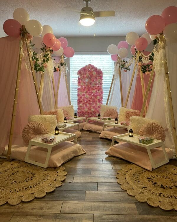 Immerse yourself in a whimsical indoor oasis with this cozy setup, perfect for a magical movie night experience. Delight in the enchanting pink and white teepee tents surrounded by plush cushions and elegant low tables, creating an intimate setting. The scene is adorned with vibrant balloons and lush floral decor, adding a splash of color and charm. An inflatable projector screen hints at a playful outdoor cinema vibe, while soft lighting casts a warm glow across decorative rugs, enhancing the inviting atmosphere. This charming indoor retreat is ideal for those seeking a whimsical yet comfortable gathering space. Keywords: cozy indoor setup, whimsical movie night ambiance, pink and white tents, floral decor, inflatable screen cinema experience.