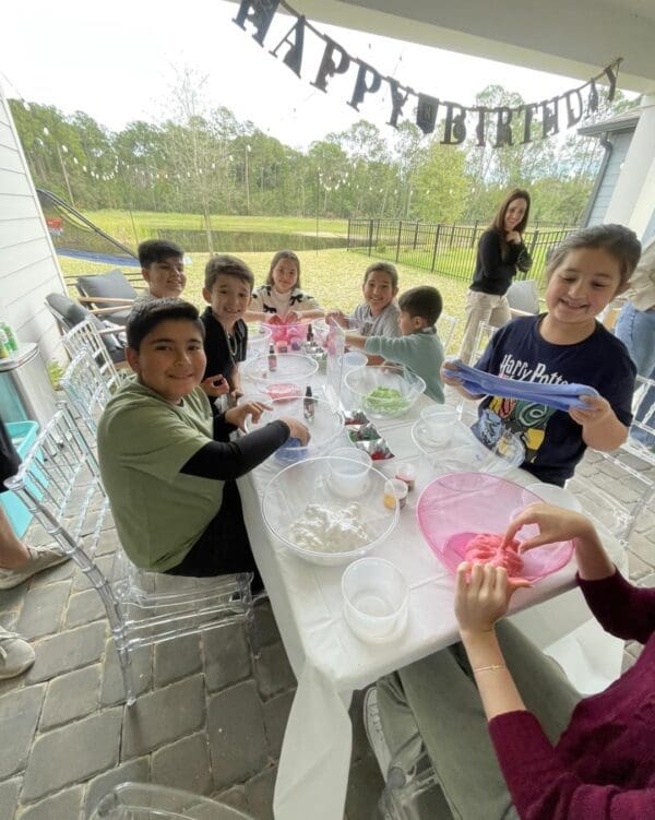Children enthusiastically gather around a table, creatively crafting vibrant slime during an exciting outdoor birthday celebration. In the background, a colorful projector illuminates the scene with vivid patterns, enhancing the festive atmosphere. The area is adorned with enchanting decorations and lush greenery, setting a picturesque backdrop for an unforgettable movie night under the stars on a large inflatable screen. This immersive experience combines DIY fun and cinematic magic in an idyllic garden setting.