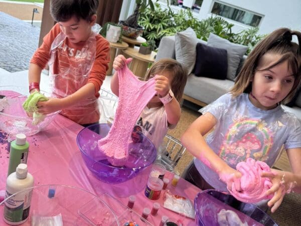 Three enthusiastic children gather around a table, fully immersed in the art of crafting vibrant, gooey slime. An outdoor projector beams vivid, enchanting visuals onto a large inflatable screen in the background, setting the stage with a mesmerizing display that enhances their creative chaos. The children's joyous laughter dances through the air, harmonizing with the soft whirring of nearby equipment to create an atmosphere brimming with fun and boundless imagination—perfect for a memorable backyard playtime experience. Keywords: kids making slime, outdoor projector fun, backyard playtime activities.