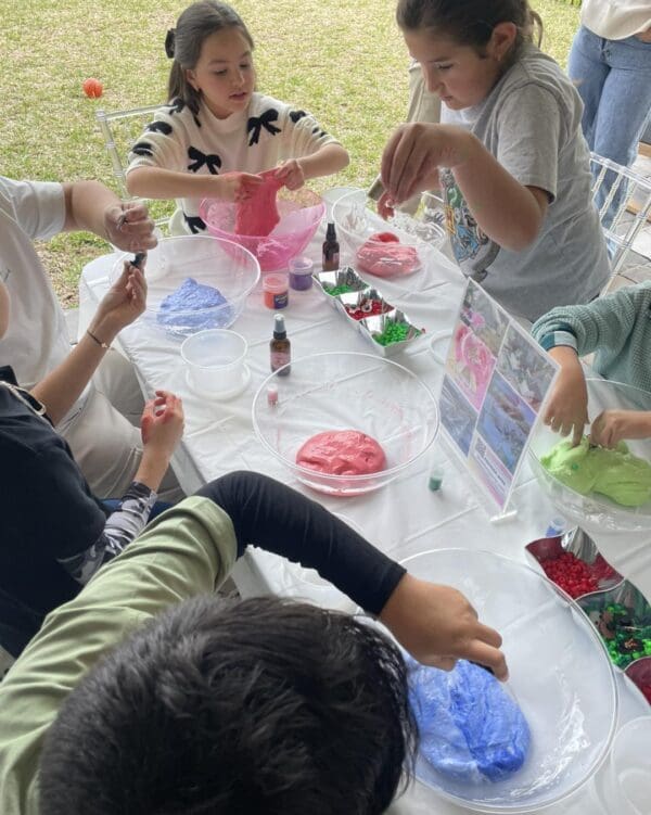 Children Enthusiastically Craft Vibrant Slime Creations for an Unforgettable Movie Night Under the Stars - A group of excited children gather around a table, their faces lit with joy as they mix and mold colorful slime using a variety of tools including bowls, bright dyes, and shimmering plastic beads. In the background, the setting sun casts a warm glow over an inflatable screen and outdoor projector poised to transform the backyard into a captivating open-air cinema experience under the twinkling night sky. This delightful scene combines creativity and magic, perfect for family bonding in this enchanting evening setup.