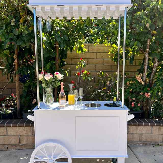 A white cart with a canopy holds a vase of flowers, champagne, orange juice, and glasses—perfect for a glamorous Mother’s Day lounge party in a garden with lush foliage and a brick wall.