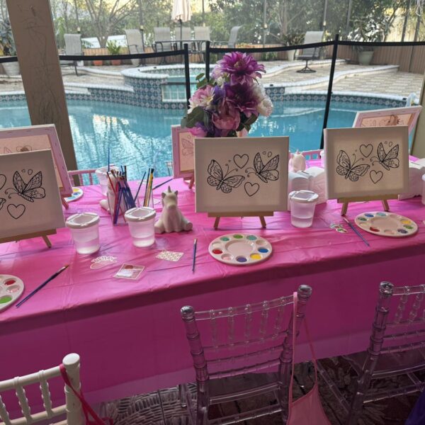 A pink-covered table by a pool is set up for a fun kids activity, featuring butterfly coloring canvases, paint palettes, brushes, and water cups ready for a creative painting session.
