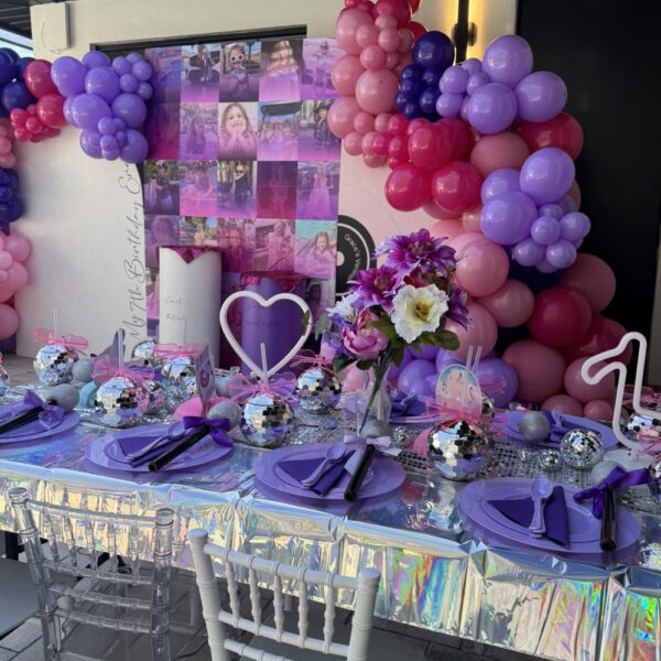 A party table with purple plates, disco ball centerpieces, and iridescent tablecloth sets a festive mood for an outdoor movie night Florida, complete with pink and purple balloons and a collage backdrop decorated with photos and flowers.