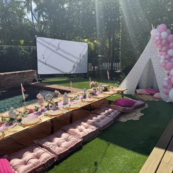 Outdoor garden setup by the pool with low picnic tables, pink cushions, a white teepee adorned with pink balloons, and a large projector screen—perfect for your next celebration from a top party shop in South Florida or Central Florida.