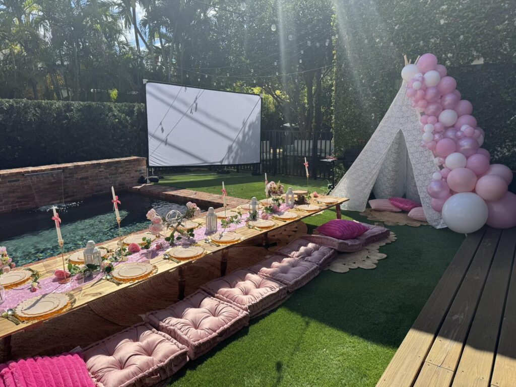 Outdoor movie night Florida vibes: a garden setup with low wooden tables, pink cushions, a decorated teepee with pink balloons, and a large projector screen by the pool.