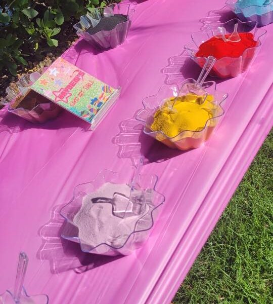 Several bowls filled with different colors of powder, each with a scoop, are arranged on a pink tablecloth outdoors next to a colorful kids activity box.