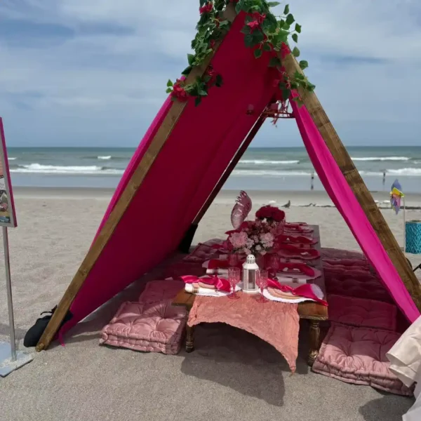 An Extra Large Teepee Rental in pink includes a low dining table and floor cushions, set up on a sandy beach with ocean views.