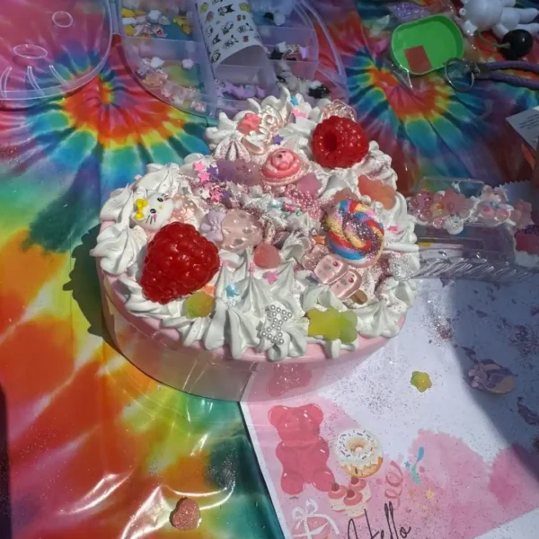 A heart-shaped cake with candies, sprinkles, and cream sits on a tie-dye tablecloth next to craft supplies, pink illustrated paper, and an Extra Large Teepee Rental set up for a cozy celebration.