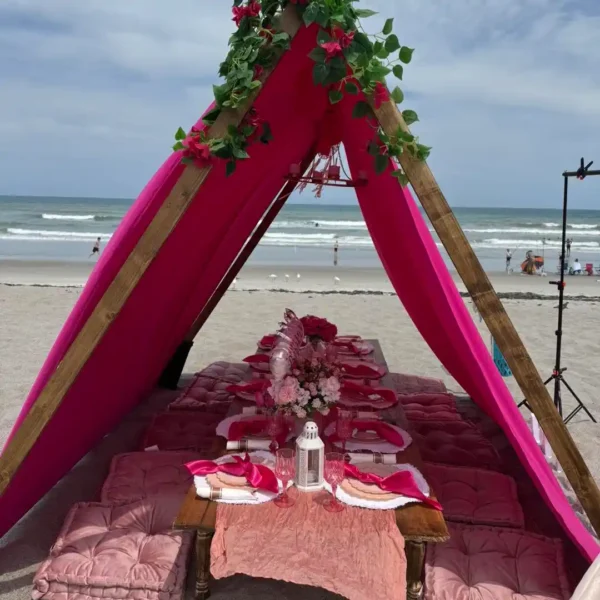 An Extra Large Teepee Rental with wooden supports covers a beach picnic setup featuring floor cushions, a low table with pink tableware, and floral decorations by the ocean.